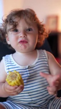 Bebek iki eliyle mısır koçanı tutuyor ve otururken onu ısırıyor, sıcak ev ortamında yemek yerken atıştırmaya odaklanıyor.