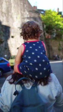 Kıvırcık saçlı çocuk şehir caddesinde yürürken omuzlarına oturmuş mavi sırt çantası ve çizgili gömleği arkadan görülüyor..