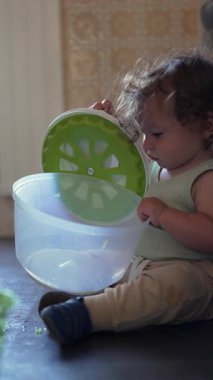 Yürüyen çocuk yerde dönen çanağı açarken tek başına mutfakta oyun oynuyordu. İçeriği merak ve ev ortamında nazik bir konsantrasyonla inceliyordu.