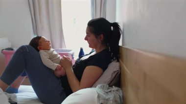 Anne ve bebek yatakta güzel bir anı paylaşıyorlar. Anne oturur, sevgiyle bebeğine bakar. Pencereden gün ışığı ve arka planda rahat pembe bir beşik..