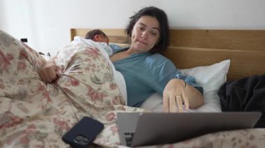 Anne yatakta, yeni doğmuş bebek göğsünde uyurken, dizüstü bilgisayar kullanarak, bakım ve rahatlamayı dengeleyerek, ev ortamında rahat bir şekilde oturarak, ebeveynlik ve aile ortamında birden fazla görev yaparak,.