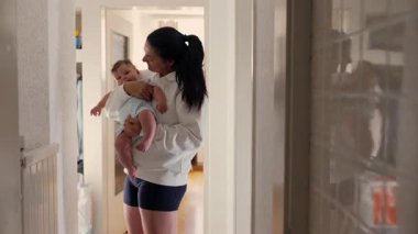 Genç anne bebeğini nazikçe kucaklıyor, koridorda dikilirken bebeğin başını öpüyor. Yansıma yakınlardaki bir aynaya yansıdı..