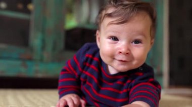 Bebek karnında yatıyor, heyecandan gülümsüyor, doğrudan kameraya bakıyor, neşeli ifade, emekleme evresi, merak, oyunculuk, ev ortamı, bebek gelişimi