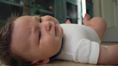 Bebek tedirgin bir ifadeyle yerde yatıyor, hafif kaşları çatık, gözler rahatlık arıyor, vücut gerilimi, uzanma ihtiyacı, dikkat çekme ihtiyacı, evde duygusal bir an.