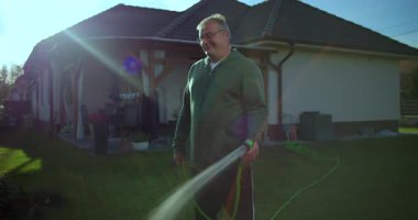 Arka planda bahçe hortumu, konut evi ve avlusu olan bahçeyi sulayan bir adam, banliyö evlerinde yeşil ve güneş ışığı olan rahat ve rahat bir an..