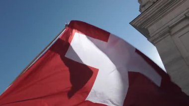 Parlak kırmızı arka plan ve beyaz haçla rüzgarda dalgalanan İsviçre bayrağı, açık mavi gökyüzüne ve kısmi şehir mimarisine karşı aşağıdan çekilmiş.