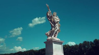 Mavi gökyüzüne karşı büyük bir haç tutan bir melek heykeli ve Roma, İtalya 'da, arka planda yemyeşil ağaçlar ve klasik Avrupa mimari atmosferi ile
