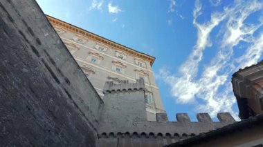 Tarihi taş tahkimatı ve Vatikan Rönesans binası. Eski savunma duvarları ve papalık mimarisinin üzerinde mavi gökyüzünde dramatik bulutlar var.