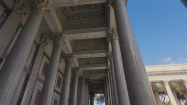 Süslü büyük harflerle süslemeli uzun taş sütunlar klasik mimaride dekore edilmiş tavanı, mavi gökyüzü ve arka planda ağaçlarla dış görünüşü destekliyor.