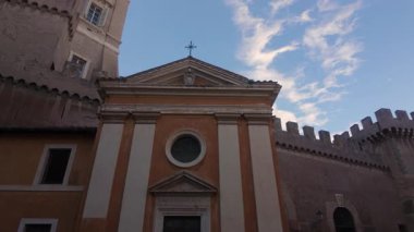 Dairesel pencereli tarihi Avrupa kilisesi cephesi ve çatıda mavi gökyüzüne karşı bulutlarla çevrili, güçlendirilmiş ortaçağ duvarları ve mimari ayrıntılarla çevrili.