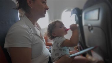 Uçaktaki çocuk, annesinin kucağında otururken saman şırıngasından içiyor, kabin içi görünür bir şekilde uçuş sırasında faaliyet kitabını tutuyor..