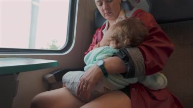 Anne, sessiz bir yolculuk sırasında tren penceresinin yanında otururken bebeğini emziriyor. Çocuk dikkatlice ve dikkatlice tutuluyor.