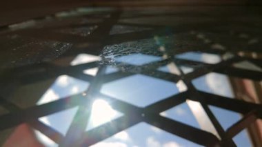 Pencere camındaki tozlu metal ızgaradan yansıyan güneş ışığı görünür mercek alevi, bulutları ve geometrik gölgeleri katmanlı perspektifte yakalıyor.