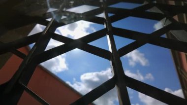 Demir ızgarayı delip geçen güneş ışığının düşük açılı görüntüsü gökyüzü ve arkaplandaki dağınık bulutlar, yıpranmış metal yapıyı ve açık alanı aydınlatıyor.