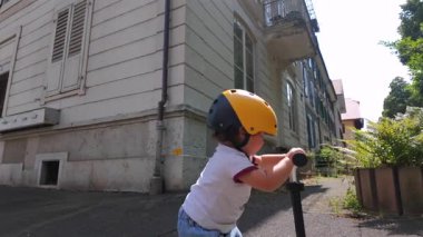 Güneşli bir günde apartman binasının dışında scooter 'la gezen bebek, kenar mahallelerdeki fabrikaların ve apartman pencerelerinin yanındaki kaldırımda süzülüyor.