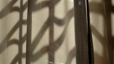 Gün ışığında geometrik pencere parmaklıkları tarafından şeffaf perdeye dökülen soyut gölge desenleri, kumaş yüzeyinde katmanlı doku ve kontrastlar oluşturur.