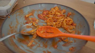 Tahta masada domates soslu bir tabak tagliatelle makarna, metal kaşık ve turuncu servis kaşığıyla servis edilir.