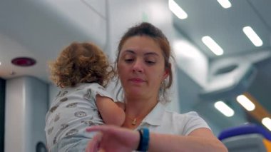 Anne, modern tren vagonunda kıvırcık saçlı ve hayvan desenli tişörtlü çocuk kucağında. Aile seyahati sırasında parlak ışıklar altında.