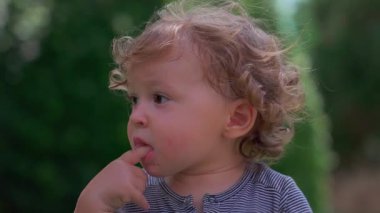 Dalgın dalgın bakan kıvırcık saçlı bir çocuk dışarı bakıyor, parmakları dudaklarında dinleniyor.