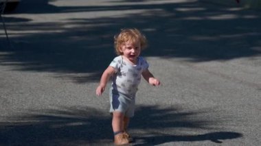 Güneşli açık havada kaldırımda koşan kıvırcık saçlı, gülümseyen çocuk yerde uzun gölgeler bırakıyor.