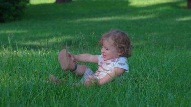 Küçük çocuk uzun yeşil çimenlerde oturuyor, kollarını kaldırıyor, neşeli bir enerjiyle, ağzı açık, sanki konuşuyor ya da şarkı söylüyor gibi, ılık yaz günü, yemyeşil bir parkta.