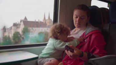 Anne akıllı telefona bakarken yürümekte olan çocuk Avrupa 'nın bulanık manzarası ve kilise kulesiyle hareket halindeki trende dikkat çekmeye çalışıyor.