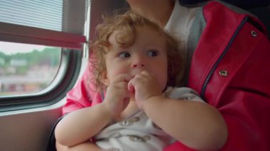 Trende annelerin kucağında oturan, gündüz vakti pencereden dışarı bakarken atıştırmalık yiyen bir çocuk..