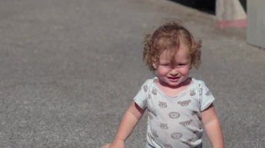 Kıvırcık saçlı çocuk güneşli bir günde dışarıda neşeli bir ifadeyle yürüyor ve elini kaldırıyor.