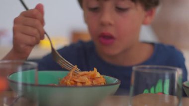 Mavi gömlekli genç yemek masasında çatallı yeşil kaseden makarna yiyor. Etrafı gözlük ve ev eşyalarıyla çevrili.