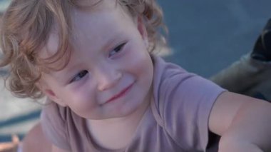 Kıvırcık saçlı çocuk biraz endişeli bir ifadeyle yana bakıyor, küçük bebek dişleri gösteriyor, ilgi arıyor ve çocukluk döneminin hassas duygularını yakalıyor.