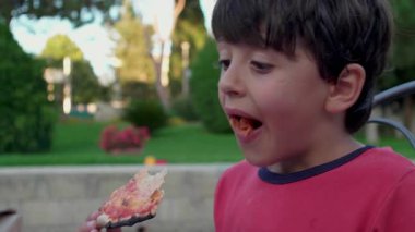 Kırmızı tişörtlü çocuk elinde bir dilim pizza tutuyordu ve dikkatlice yukarı bakıyordu. Güneşli bir günde bahçede yemek yerken bir an durakladı.