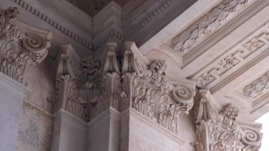 Corinthian başkentlerinin mermerden yapılmış köşeleri, akanthus yaprakları, voltajlar ve dekoratif kıvrımlarla zenginleştirilmiş, karmaşık desenli tavanın altında.