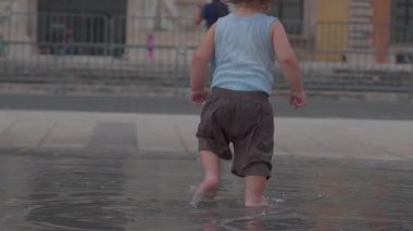 Kıvırcık sarı saçlı çocuk şehir çeşmesindeki sığ sularda yalın ayak yürüyor, arkasından bakılıyor, halka açık meydanda tasasız enerji saçıyor.