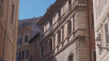 Açık mavi gökyüzünün altında dar bir sokakta kemerli camları, taş cepheleri ve klasik balkonları olan tarihi Roma binaları geleneksel Avrupa mimarisini gözler önüne seriyor.