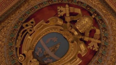 Üzerinde taç, çapraz anahtarlar, defne çelengi ve kraliyet sembolleri olan mavi bir kalkan bulunan süslü kasalı tavan madalyonu içindeki altın papalık armasının yakın ayrıntıları.
