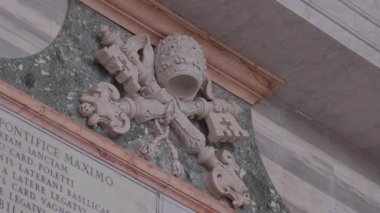 Papalık arması mermere oyulmuş taç ve çapraz anahtarlar, yeşil mermer panel üzerine monte edilmiş ve tarihi bazilika içinde Latince yazılar