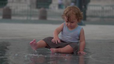 Islak yüzeyde oturan çocuk, sığ su ve arka plan yapıları olan şehir meydanında yan tarafa bakıyor.