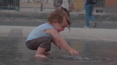 Kıvırcık sarı saçlı bebek şehir çeşmesindeki sığ suyun üzerinde yalın ayak, dalgalara doğru uzanan, bulanık yayalar ve şehrin arka planında