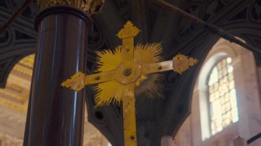 Merkezi güneş patlaması detayı içeren altın işlemeli haç, tarihi bazilika içinde mermer sütun önünde kemerli pencere ve yukarıda karmaşık tavan