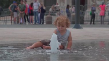 Küçük çocuk şehir meydanında yer seviyesindeki çeşmenin yanında oturuyor, dalgaların içinde el çırpıyor ve suyu yakından izliyor, ferahlatıcı yaz anının tadını çıkarıyor.