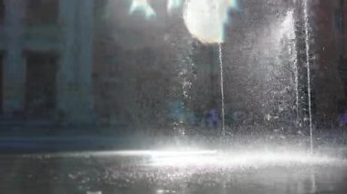 Şehir meydanında fıskiye fışkırıyor, güneş ışığı su damlacıklarını kırıyor şehir meydanında mimari arka planda parlak sisli bir atmosfer yaratıyor.