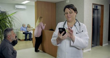 Hastanenin resepsiyonunda telefonla gülümseyen kadın doktor hastalar beklerken ve çalışanlar arka planda yürürken.