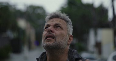 Yukarı bakan orta yaşlı bir adam, yağmurda ellerini kaldırmış, duygusal sorgulama ya da ruhani bir katman.