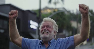 Mavi gömlekli yaşlı adam kollarını kaldırdı ve neşeli bir ifade takındı. Yağmurun altında coşkuyla gülümsüyordu.