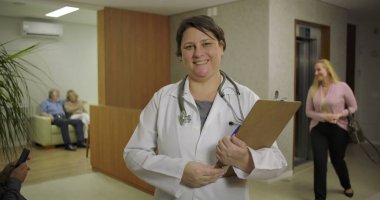 Hastanenin lobisinde hastalar ve resepsiyonistle birlikte asansörün yanında pano tutarken gülümseyen doktor.