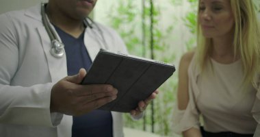 Klinik ortamdaki orta yaşlı kadın hastalara tıbbi verileri gösteren bir tablet tutan doktor, açık iletişim ve modern bakım araçlarını vurguladı