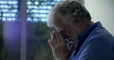 Pencerenin yanında duran mavi gömlekli yaşlı adam, gözleri kapalı ve elleri derin duygusal dualarla ya da hüzünlü yansımalarla birleşmiş.