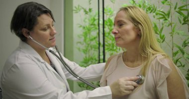 Doktor orta yaşlı kadın hastaların kalp atışlarını dinlemek için steteskop kullanıyor. Klinikte yeşil bitki duvarı arka planına sahip tıbbi muayene sırasında.