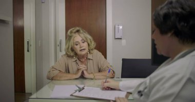 Kıdemli kadın, tıbbi büro danışmanlığında bayan doktorla konuşurken kaygısını dile getiriyor. Masanın karşısında oturuyor. Evrak işleri ve bilgisayarla arka grupta görülüyor.