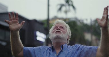 Kolları geniş ve yüzü göğe yükselmiş yaşlı bir adam, ruhsal rahatlama, minnettarlık veya duygusal teslimiyet ifadesiyle yağmuru kucaklıyor.